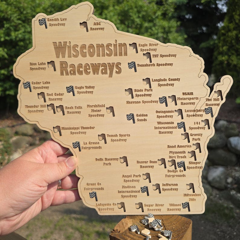 May include: Wooden map of Wisconsin with the words "Wisconsin Raceways" and the names of various speedways. The map is light brown with dark brown text and small checkered flag icons. A hand is holding the map.