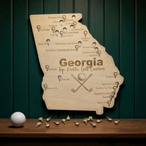May include: A wooden map of Georgia with the text "Georgia Top Public Golf Courses" engraved. The map features golf course markers and a golf ball with crossed clubs. A golf ball and markers are on the wooden surface.