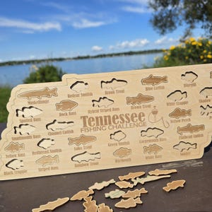 May include: A light-coloured wooden board shaped like the state of Tennessee, featuring cutouts for various fish species. The text "Tennessee FISHING CHALLENGE" is engraved in the centre, surrounded by labels for fish like Muskellunge and Bluegill.