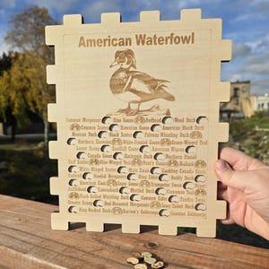 May include: A wooden puzzle with the words "American Waterfowl" and an engraved Wood Duck illustration. The puzzle has cutouts and labels for various North American waterfowl species, including the Common Merganser and the Blue Goose. The puzzle is being held by a hand.