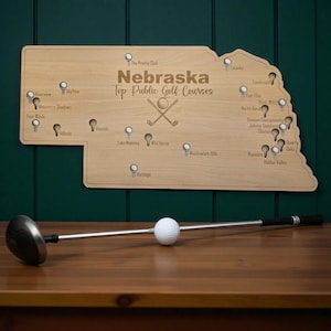 May include: A wooden map of Nebraska with the text "Nebraska Top Public Golf Courses" and golf course pin markers. A golf club and a golf ball are on a wooden surface in front of the map.