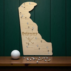 May include: A wooden map of Delaware with golf course locations marked. The map is light brown and shaped like the state. The text "Delaware Top Public Golf Courses" is displayed. A golf ball and tees are also present.