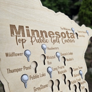 May include: A wooden map of Minnesota with the text "Minnesota Top Public Golf Courses" engraved. The map features golf ball-shaped markers and the names of golf courses. The wood has a light, natural finish.