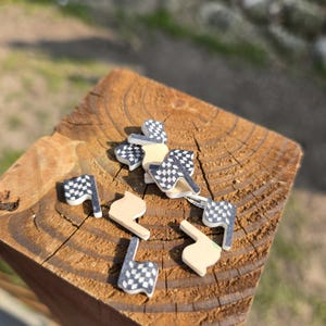 May include: A collection of small, wooden racing-themed embellishments. The assortment includes checkered flags in black and white, and beige-colored shapes. The items are scattered on a weathered, brown wooden surface.