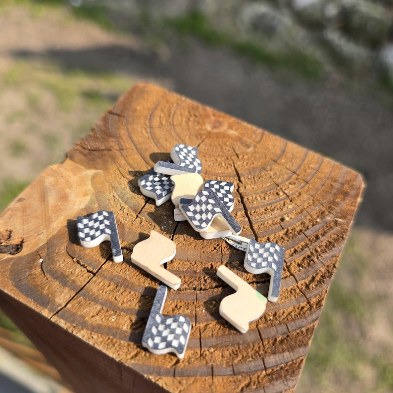 May include: A collection of small, wooden racing-themed embellishments. The assortment includes checkered flags in black and white, and beige-colored shapes. The items are scattered on a weathered, brown wooden surface.