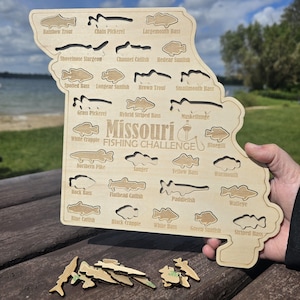 May include: A wooden jigsaw puzzle shaped like Missouri, with fish outlines including Rainbow Trout and Largemouth Bass. The puzzle is labelled "Missouri Fishing Challenge" and has several fish-shaped pieces.