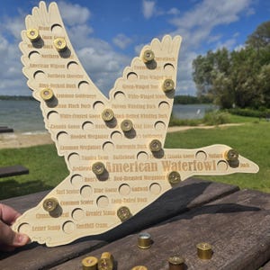 May include: A light-colored wooden display board shaped like a flying bird, featuring the text American Waterfowl and numerous circular cutouts labeled with bird species names. Several brass shotgun shell casings are inserted into the holes.