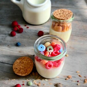 May include: Two glass jars with lids filled with a white candle and colourful cereal pieces. The jars are on a wooden surface with raspberries and blueberries scattered around.