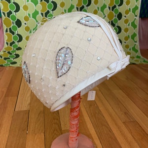 May include: Cream-colored vintage hat with a delicate net overlay and decorative sequins. The hat features leaf-shaped embellishments and is displayed on a pink stand. A white ribbon accents the brim, adding a touch of elegance.