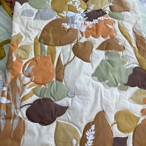 May include: A quilted fabric with a leaf pattern in shades of brown, green, and gold on a cream background. White floral accents are interspersed throughout the design. The fabric is likely for home decor or crafting.