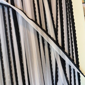 May include: Close-up of a sheer fabric with vertical black stripes and a scalloped black trim. The fabric is folded, creating a pleated effect. The material appears lightweight and airy, suitable for curtains or decorative purposes.