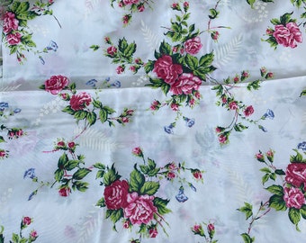 May include: A white fabric featuring a repeating floral pattern of deep pink roses, green leaves, and small blue bell-shaped flowers. The design includes delicate white fern-like foliage accents scattered across the smooth surface.