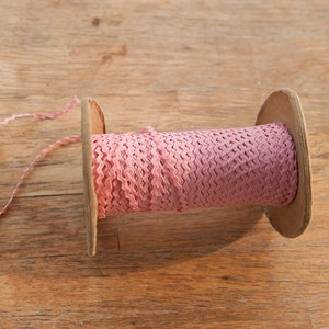 May include: A spool of pink ric rac ribbon sits on a wooden surface. The ribbon is tightly wound around a cardboard spool. A length of the ribbon extends from the spool, showing its zig-zag edge. The ribbon and spool are in a close-up shot.