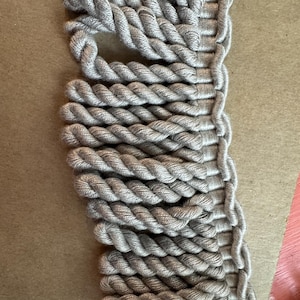 May include: Close-up of a section of grey rope trim. The trim is made of tightly twisted rope, creating a textured surface. The rope is a neutral grey colour, suitable for various craft projects or home decor.