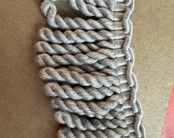 May include: Close-up of a section of gray rope trim. The trim is made of tightly twisted rope, creating a textured surface. The rope is a neutral gray color, suitable for various craft projects or home decor.