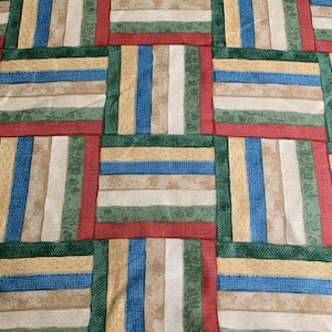 May include: A patchwork quilt featuring a repeating pattern of rectangular blocks. Each block is composed of vertical stripes in various colors, including red, green, blue, beige, and white. The quilt has a textured appearance.