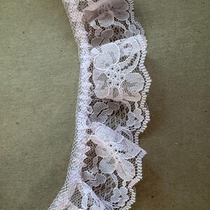 May include: A close-up of a lavender-coloured lace trim with a ruffled edge. The lace has a detailed floral design and a scalloped border. Suitable for sewing and crafts, this trim adds a vintage aesthetic to projects.
