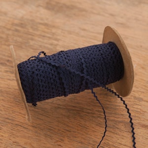 May include: A spool of navy blue ric rac trim. The trim is tightly wound around a cardboard spool, with a small length of the trim extending out. The trim has a zig-zag edge. The background is a light brown wooden surface.