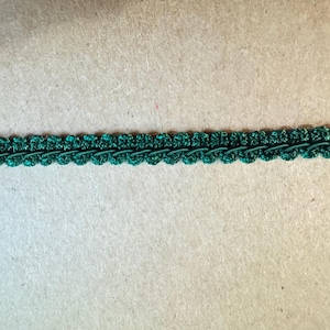 May include: A length of dark green decorative trim with a scalloped edge. The trim has a textured, woven appearance and is likely used for embellishing clothing, crafts, or home decor projects.