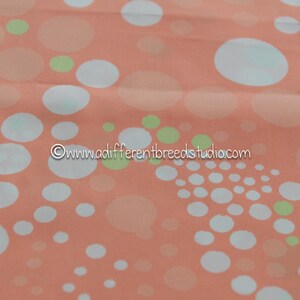 May include: Peach-colored fabric with a repeating pattern of white, light green, and peach-colored circles of varying sizes. The fabric has a soft, flowing appearance, suitable for apparel or home decor.