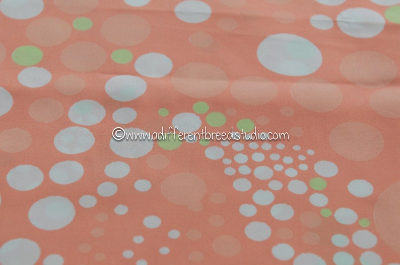 May include: Peach-colored fabric with a repeating pattern of white, light green, and peach-colored circles of varying sizes. The fabric has a soft, flowing appearance, suitable for apparel or home decor.