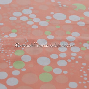 May include: Peach-colored fabric with a pattern of white and light green circles of varying sizes. The circles are scattered across the fabric, creating a playful and modern design. The text "www.adifferentbreedsstudio.com" is in the image.