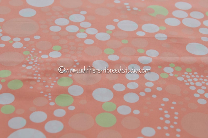 May include: Peach-colored fabric with a pattern of white and light green circles of varying sizes. The circles are scattered across the fabric, creating a playful and modern design. The text "www.adifferentbreedsstudio.com" is in the image.