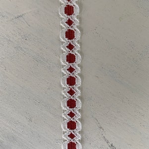 May include: A decorative trim featuring a repeating pattern of white openwork ovals and diamonds, framing a solid red center. The trim is displayed against a textured, off-white surface, showcasing its detailed design.