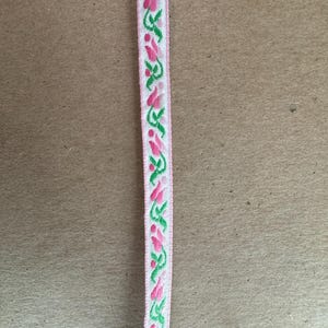 May include: A white ribbon with a pink border and a repeating pattern of pink flowers and green leaves. The ribbon is likely used for crafts, sewing, or gift wrapping.