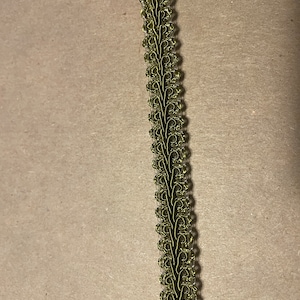 May include: A strip of decorative trim with a repeating floral design. The trim features a dark green base with a metallic gold-colored overlay, creating a textured and ornate appearance. The trim is likely used for embellishing clothing or craft projects.