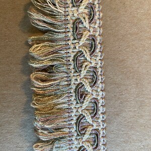 May include: Decorative fringe trim with a woven design and looped accents. The trim features a mix of beige, green, and brown threads, creating a textured and detailed appearance. The fringe adds a soft, flowing element to the design.