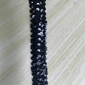 May include: A strip of black sequins, arranged in a vertical line. The sequins are round and shiny, reflecting light. The background is a light-colored wooden surface.