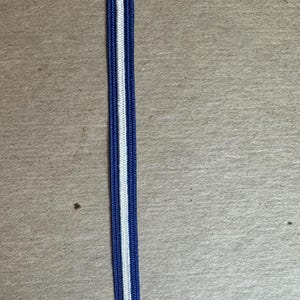 May include: A length of blue and white striped elastic ribbon. The ribbon has a central white stripe bordered by two blue stripes. The ribbon is likely used for sewing or crafting projects. The background is a neutral tan color.