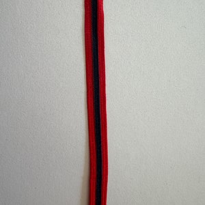 Peut inclure: Un ruban rayé rouge et noir. Le ruban présente une bande noire centrale bordée de rayures rouges de chaque côté. Le ruban est orienté verticalement sur un fond uni blanc cassé.