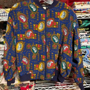 May include: A blue bomber jacket with a repeating pattern of framed classical portraits in red, yellow, and green. The jacket has a navy blue collar, cuffs, and waistband, and a full-length zipper. The background is a cluttered shelf.