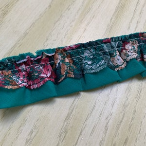 May include: Close-up of a strip of decorative trim. The trim features a teal base with a ruffled edge. The ruffled edge is adorned with floral lace in shades of burgundy, brown, and white. The trim is sewn with white thread.