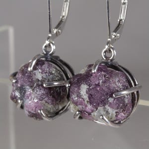 May include: A pair of drop earrings featuring raw, deep purple and white mineral stones held in place by silver-toned metal prongs. Each earring is suspended from a silver-toned lever-back closure, showcasing the natural, irregular texture of the stones.