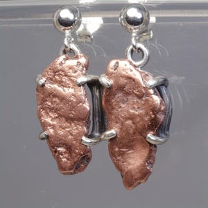 May include: A pair of drop earrings featuring raw, textured copper nuggets secured in silver-toned metal prong settings. Each earring hangs from a polished silver-toned ball stud post, showcasing the contrast between the metallic copper and silver.