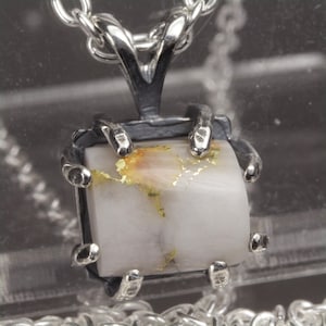 May include: A silver pendant necklace featuring a square, white stone with gold flecks. The stone is held in place by a silver setting with claw-like prongs. The chain is also silver, creating a cohesive jewelry piece.
