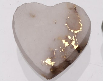 Può includere: Una pietra a forma di cuore con una superficie liscia color bianco sporco, caratterizzata da texture cristalline naturali e irregolari accenti metallici color oro incastonati nella superficie, creando un netto contrasto.