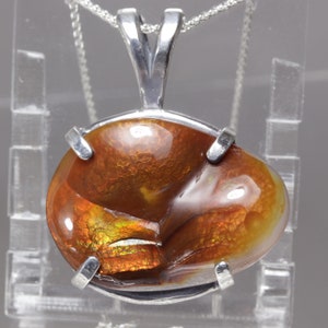 May include: A polished, oval-shaped fire agate gemstone pendant set in a silver prong setting. The stone displays deep reddish-brown tones with iridescent flashes of orange and gold. The pendant hangs from a thin silver chain, showcasing a smooth, reflective surface with a distinct internal play of color and natural layered texture.