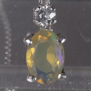 May include: A close-up of a pendant featuring an oval-cut, iridescent gemstone. The gemstone is held in place by four silver prongs and is connected to a small, clear, round stone at the top.