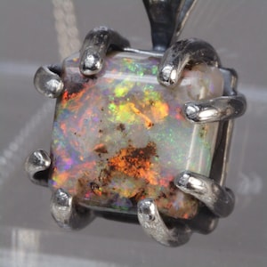 May include: A square-cut opal pendant set in a silver claw setting. The opal displays a vibrant play of color with flashes of red, orange, green, and blue against a milky white base. The metal setting features six prongs that secure the stone.