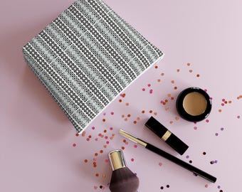 May include: A black and white patterned makeup bag with a makeup brush, lipstick, and a small container of makeup on a pink surface with pink and gold confetti.
