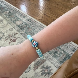 May include: A blue and white beaded bracelet with a pink and blue flower charm. The bracelet is made of polymer clay beads and has a gold accent.