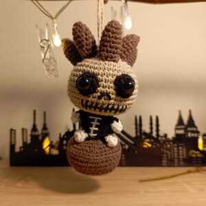 Könnte beinhalten: Eine gehäkelte Skelettpuppe aus braunem und beigefarbenem Garn, die ein schwarz-weißes Skelettkostüm trägt. Die Puppe hat große schwarze Augen und sitzt auf einer braunen gehäkelten Kugel.