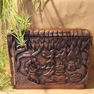 May include: A dark brown wooden box with detailed carvings of a village scene, including figures, trees, and a rabbit. The rectangular box has a textured surface, demonstrating skilled craftsmanship and a rustic aesthetic.
