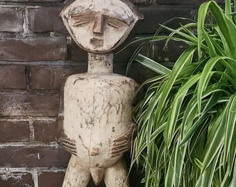 Può includere: Una statua in legno scolorito, bianco sporco, di una figura umana con una grande testa e tratti stilizzati. La figura ha un busto arrotondato e sta in piedi su due gambe. La statua è posta contro un muro di mattoni e piante verdi.