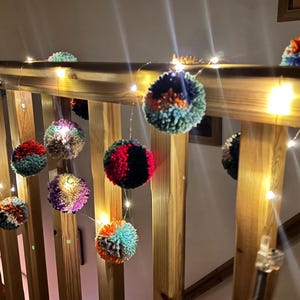 May include: A string of decorative fairy lights featuring multi-coloured yarn pom-poms in shades of red, blue, green, and purple. The small, round, fuzzy pom-poms are interspersed with warm white LED lights along a thin, flexible wire cable.