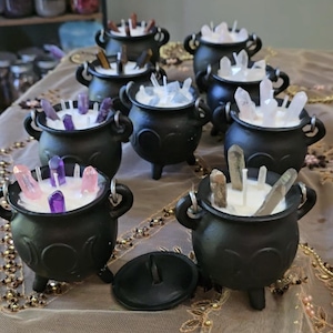 May include: A collection of black cast iron-style mini cauldrons filled with white wax and embedded with various raw crystal points. Each cauldron features three small legs, two side handles, and a triple moon symbol embossed on the front.